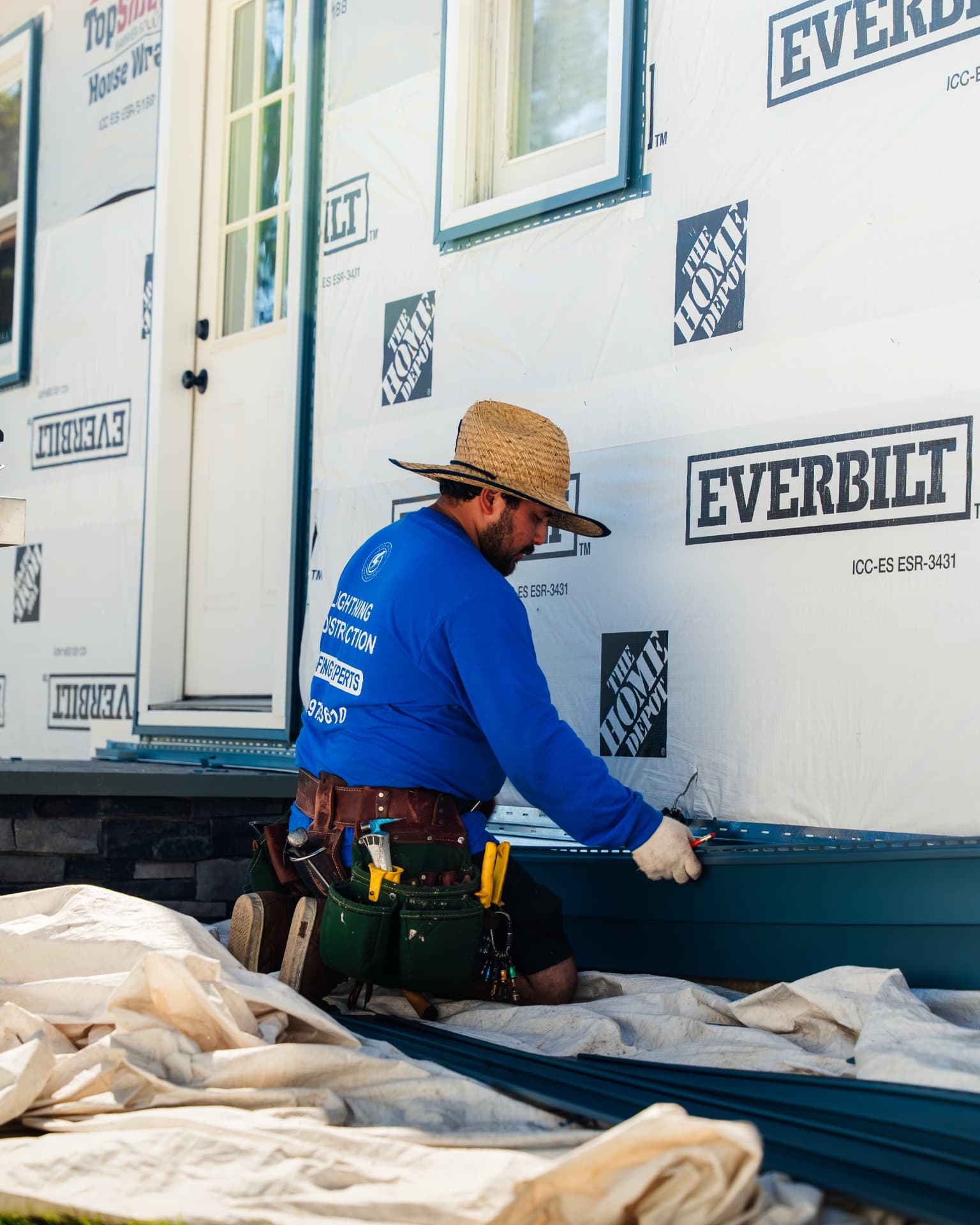 Construction worker in a Lightning Construction shirt kneeling on a tarp installing dark siding onto a house wrapped in house wrap